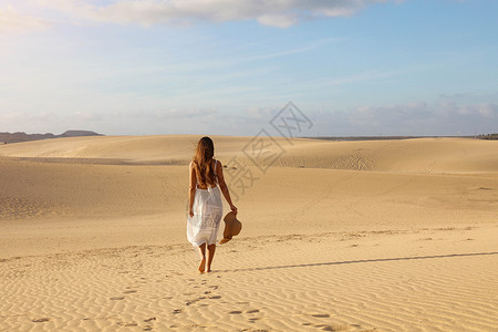 日落时在沙漠沙丘行走的年轻美女身着白裙子 女孩在科拉莱乔杜纳斯金沙上行走背景图片