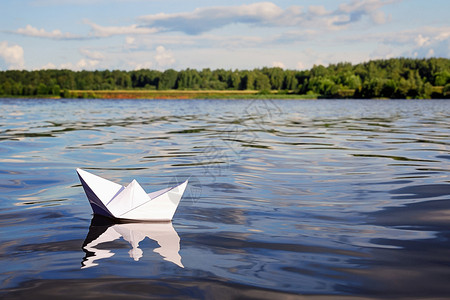 小纸船漂浮在蓝色平静的河水 绿色的森林 蓝天上 地平线上有淡淡的夏日云彩 自由 冒险 梦想和旅行概念背景