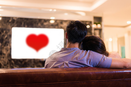 亚洲年轻情侣在沙发上看电视 夫妻和放松的概念 假期和假期的概念 夜间约会和蜜月主题 后视图 白屏电视中的红心背景图片