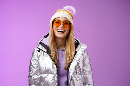 穿着银色闪亮夹克帽子太阳镜的时尚友善 魅力十足的金发女性准备学习单板滑雪 笑着开心地玩雪地休闲度假胜地 站在紫色背景下 学生 冬背景图片