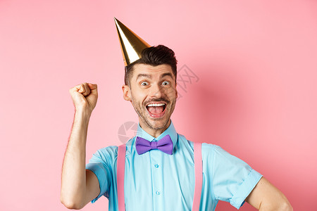 假期和庆祝活动的概念 生日快乐的男孩 留着胡子 戴着领结 戴着派对圆锥帽和节日服装 举手大喊是 吊带裤 男人背景