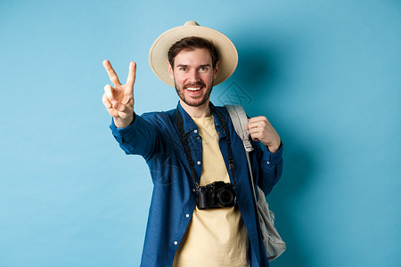 在暑假时展示和平标志 在阳光下享受热带节日 戴草帽 拿着背包和照相机 蓝背景的英俊正面男子观光客 工作室 大胡子背景图片