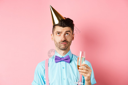 假期和庆祝活动的概念 脾气暴躁的家伙戴着生日派对帽 拿着一杯香槟 用怀疑的脸抬头看着粉红色的背景 时尚 有趣的背景图片