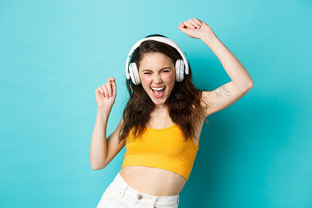 穿着夏装的年轻女性听音乐 戴着耳机 沿着最喜欢的歌曲唱歌 戴着耳机跳舞 站在蓝色背景下 喜悦背景图片