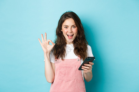 电话的标志兴奋和高兴的年轻女性的形象在使用智能手机应用程序 使用手机在线购物并显示 OK 标志后说是 站在蓝色背景上背景