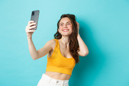 美丽的现代女孩在度假时自拍 穿着夏装摆姿势 看着智能手机相机 站在蓝色背景下 女士 广告背景图片