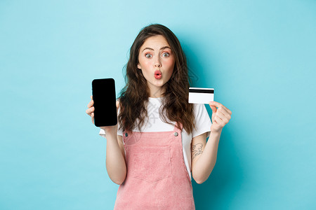 年轻美女的画像展示塑料信用卡和空的智能手机屏幕 看起来既有趣又感兴趣 展示有趣的应用程序 站在蓝色背景上背景图片