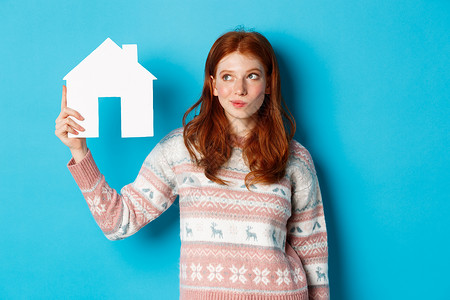房地产概念 深思熟虑的红发女孩形象 展示纸房模式和思维 寻找家或公寓 站在蓝色背景下站立 美丽的 微笑背景图片