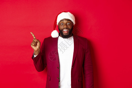 圣诞 派对和节假日概念 黑人用左手指着手指笑 展示标志 站在红色背景上 兴奋和欢乐的黑人男子 胡须 快乐背景图片