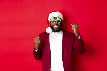 圣诞快乐 开心的黑人男子 戴着有趣的派对眼镜和圣塔帽 笑笑喜悦 庆祝冬季节日 站在红色背景上 横幅 成人背景图片