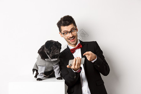 快乐的狗主人在手机上展示宠物的东西 男人和小狗穿着高雅服装 站在白色背景旁站立 新年 博主背景图片