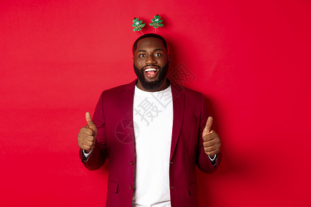 圣诞快乐 非洲男人欢庆新年快乐 穿着有趣的派对头带 举起拇指 像和赞美某样 站在红后春光前 成人 商业背景图片