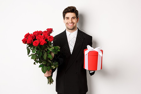 假期 关系和庆祝活动的概念 穿着黑色西装的帅气男友 拿着一束红玫瑰和一份礼物 站在白色背景上 祝圣诞快乐 大胡子 花朵背景图片