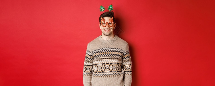 穿着派对眼镜和冬季毛衣的英俊长胡子男子的肖像 笑笑喜悦 庆祝新年 祝愿圣诞快乐 站在红色背景之上 男生 圣诞节背景图片