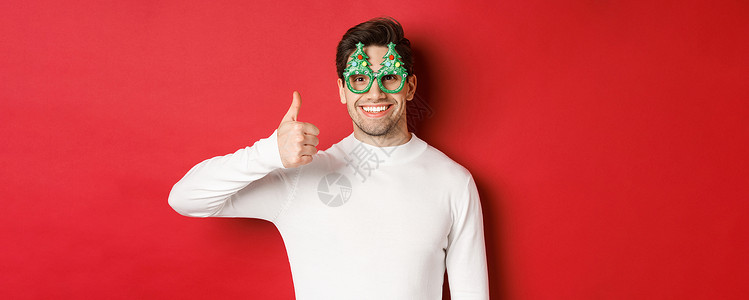 圣诞 冬季节假日和庆祝活动的概念 穿着派对眼镜和白色毛衣 举起拇指 推荐新年宣传的英俊微笑者近身露脸 圣诞节 庆典背景图片