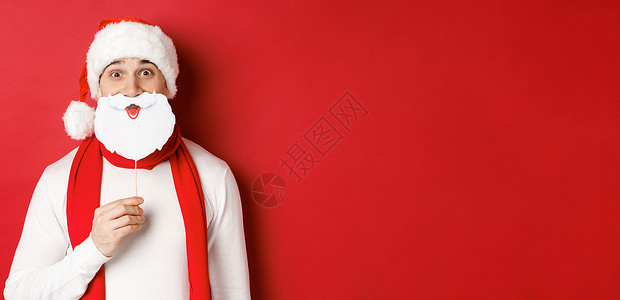 圣诞节 冬季假日和庆祝活动的概念 穿着圣塔帽 戴着胡子面具 享受新年晚会 站在红色背景上站立的滑稽男子肖像 广告 快乐背景图片