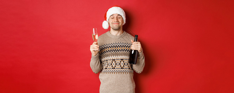寒假 圣诞节和庆祝活动的概念 戴着圣诞帽和毛衣 在新年喝酒 拿着一瓶香槟和满满的玻璃杯 红色背景的快乐微笑男人的形象 派对 黑发背景图片