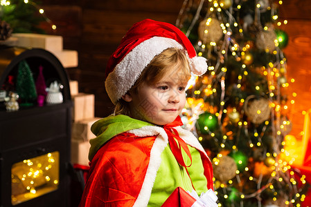 圣诞树背景下戴着圣诞老人帽子的快乐小孩 圣诞节的时候 男孩可爱的孩子快乐的心情在圣诞树附近玩耍 壁炉附近的寒假礼物背景图片