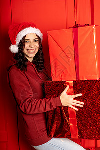 戴着圣诞老人帽子的年轻女子拿着大礼物 站在红墙附近 戴着圣诞帽的快乐女性带着一盒礼物 假期 礼物和好心情的概念 庆典 盒子背景图片