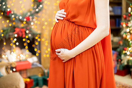 圣诞福利最好的圣诞礼物 身穿红色连衣裙的孕妇在模糊的圣诞客厅背景下抱住她的肚子 特写没有脸 女孩 母性背景