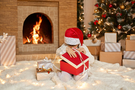 戴着圣诞帽的小女孩打开礼品盒 看着里面 穿着白色套头衫和红色圣诞老人帽 坐在节日客厅的地板上 背景是壁炉和圣诞树背景图片