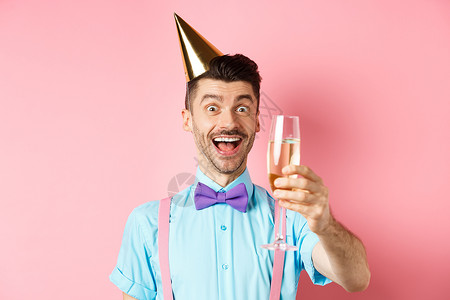 欢庆节和庆祝概念 兴奋的生日男孩 举杯香槟 做吐司或演讲 站在党帽上 享受粉红背景的乐趣 男人 吊带裤背景图片