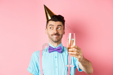 欢乐的年轻男子在派对帽上庆祝生日 想着演讲 举杯香马普涅烤面包 粉红背景 掌声 笑 快乐 手背景图片