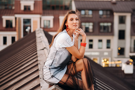 坐在屋顶上晚上 一个女孩坐在城里一所房子的屋顶上 穿着衣服和运动鞋的模特儿肖像 表情 城市屋顶背景