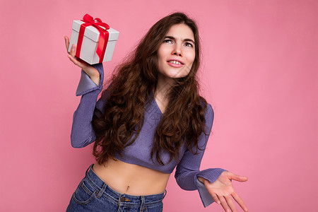 紫色上衣照片中 迷人 情绪化的黑发卷发年轻女性被隔离在粉红色背景墙上 身穿紫色衬衫 手上拿着带红丝带的白色礼盒背景