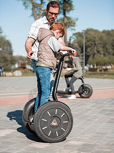 小摩托车父亲教儿子在公园骑迷你滑板背景