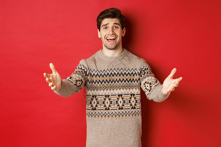 喜悦和受人欢迎的英俊男子穿着圣诞节毛衣 伸出双手迎接新年晚会的嘉宾 邀请来庆祝 站在红背景上站立 工作室 庆典背景图片
