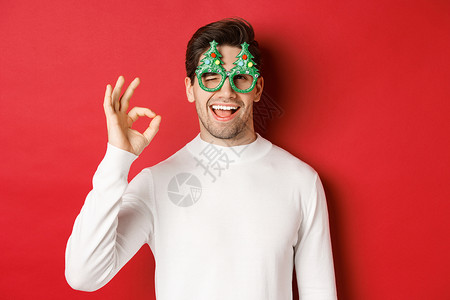 穿着政党眼镜和白色毛衣 展示好牌 眨眼 祝新年快乐 站立在红色背景之上的英俊帅气的男人肖像 大胡子 时尚背景图片