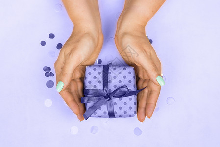 2022 年左右 女人的手上涂着蓝色柔和的美甲 手里拿着紫色背景上带蝴蝶结的礼物 节日 礼物 惊喜 生日 新年和圣诞节的概念 展背景图片