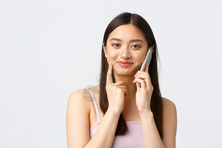 美丽 时尚和人们的情感概念 特写 美丽的亚洲女孩在电话中看起来很感兴趣 在手机上聊天 微笑着感兴趣 白色背景背景图片