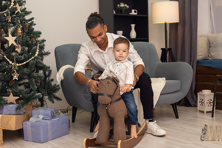 黑白父亲素材一位年轻的父亲坐在灰色沙发上 在家里把他的小儿子骑在棕色的摇马上背景