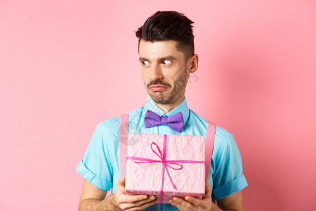 休假日和庆祝的概念 惊吓的家伙一边盯着一旁的表情 拿着小礼物盒 站在粉红背景上 乐趣背景