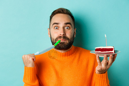 喜笑的成年男子在庆祝生日时贴近他 用蜡烛 吹舞派对节奏和欢乐 站在浅蓝背景上举着白蛋糕和蜡烛 毛衣 黑发背景图片