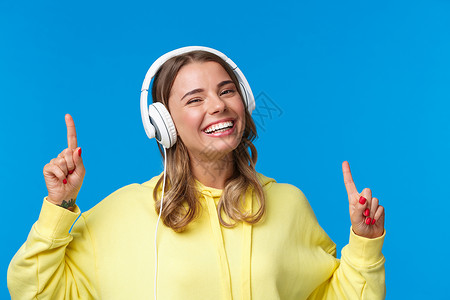 音乐 生活方式和青年概念 快乐微笑的金发女孩戴着耳机听歌 跳舞和移动手指节奏的特写 在蓝色背景中快乐地站立背景图片