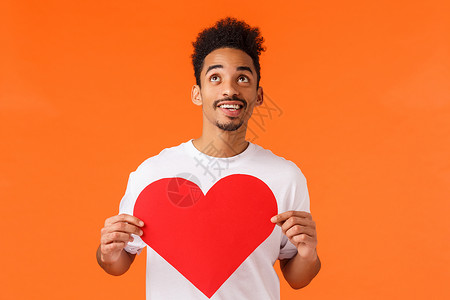梦幻浪漫的非洲裔美国男友感到鼓舞和兴奋 拿着情人节卡片 抬头微笑 寻找爱情 表达真情或同情 橙色背景背景