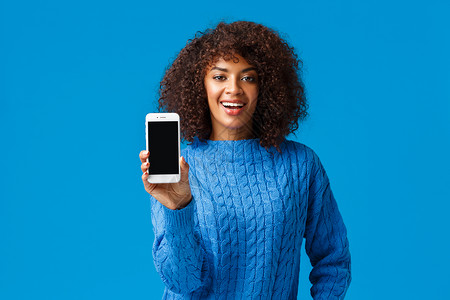 看我的成绩 快乐而自豪 漂亮的非洲裔美国女性浏览约会应用程序 展示智能手机屏幕摄像头 看看 站在蓝色背景中的冬季毛衣背景图片