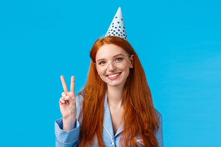 变装派对欢乐的红发欧洲女性 戴白日帽子显示和平标志 穿夜装 庆祝生日 在蓝背景上笑着 BLEWAKYT背景