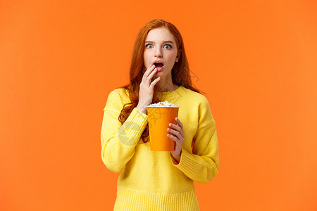 震惊和感兴趣 娱乐的红发女孩在大屏幕上观看精彩的新电影 吃爆米花 张开嘴喘着粗气 惊奇的电影情节 橙色背景背景图片