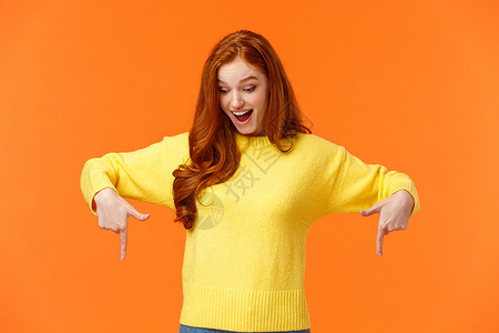 穿着黄色毛衣的可爱红发女人好奇而兴奋 逗乐了 手指向下 看着底部 微笑着 结账产品促销 寒假活动 橙色背景背景图片