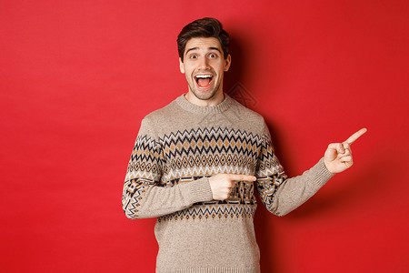 圣诞节庆祝 冬季假日和生活方式的概念 穿着Xma毛衣的帅哥指对手指 笑笑惊讶 在红色背景下展示新年宣战活动 笑声 快乐的 工作室背景