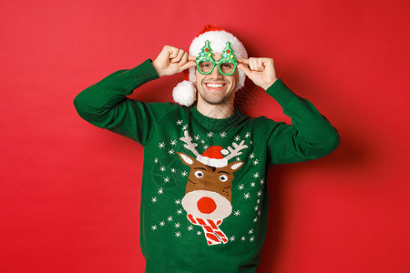 穿着圣塔帽 派对眼镜和毛衣 庆祝新年节日 站在红色背景下站立的有魅力的笑脸男子肖像 快乐的 庆典背景图片