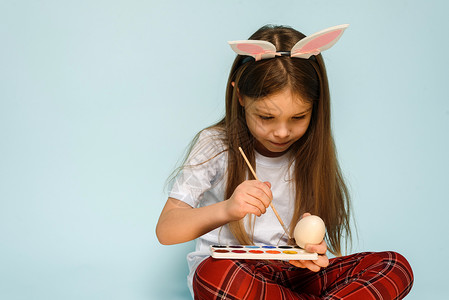 画画的兔子兔子耳朵里的一个小女孩 在工作室的蓝色背景上为复活节画彩蛋 快乐的 乐趣背景