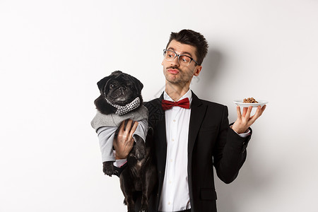 穿着漂亮西装的帅气年轻狗主 拿着可爱的黑瓜和动物食物盘子 站在白色背景上 庆典 行动背景图片