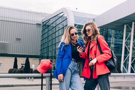 两个快乐的女孩在机场使用智能电话检查飞行或在线登机 同时携带行李 空中旅行 暑假等活动 蓝色的 背包背景图片