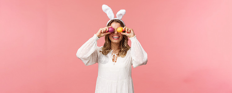 彩蛋中的女孩欢庆复活节的兔子耳朵和白裙子中的可爱 笑容温柔的女子 将彩蛋装在眼睛上 微笑 粉红背景等美人画成像 美丽 夏天背景