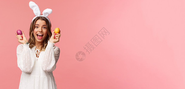 跳舞的兔子假期 春天和聚会的概念 兴奋迷人的年轻女性穿着兔耳和白色派对礼服庆祝复活节的肖像 用两个彩蛋跳舞 粉红色背景 成人 店铺背景